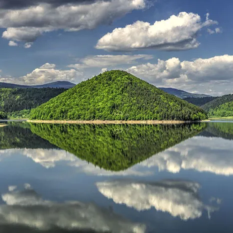 Summer landscape showing a calm lake with green forest hills in the background. Blue skz with fluffy clouds, mirrored in the surface of the lake.